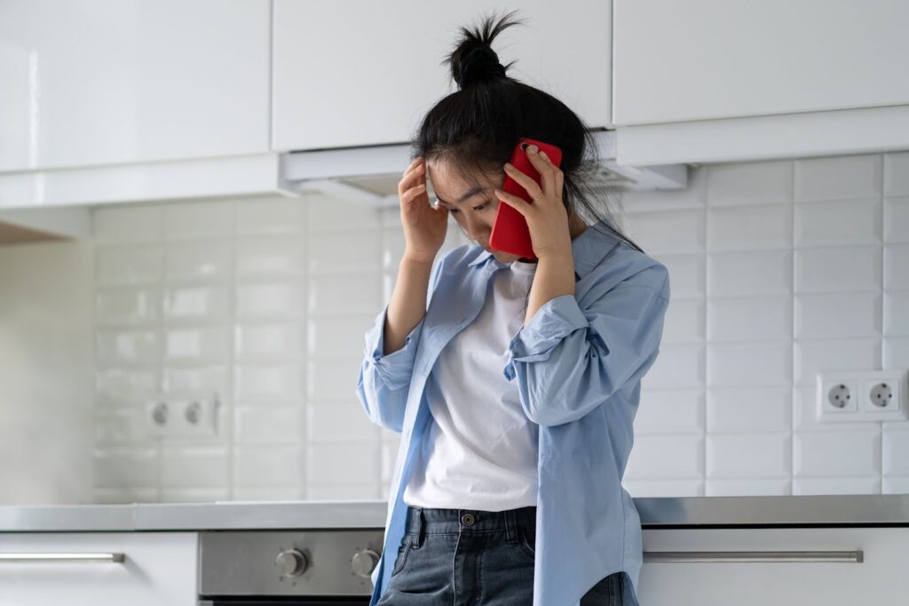 A woman stands in a kitchen holding a red phone, coordinating disaster cleanup and restoration efforts.