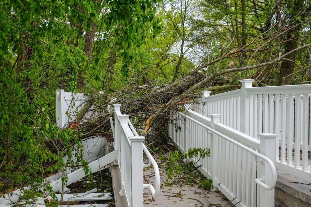 A fallen tree rests against a white railing near a house, indicating wind damage to the roof and ongoing repair efforts.